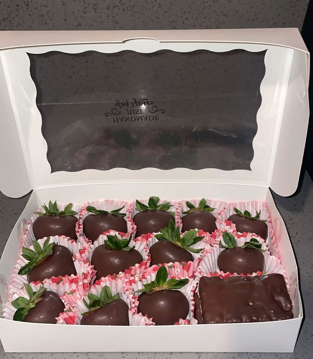A dozen classic chocolate-covered strawberries with decorative pink floral-patterned paper liners and a Rice Krispie bar in a white bakery box.
