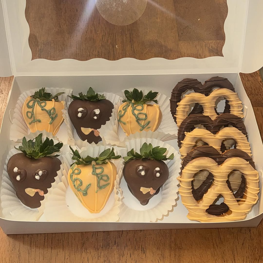 Halloween-themed box of dark chocolate and orange strawberries with playful monster faces and teal drizzle, plus matching decorated pretzels.