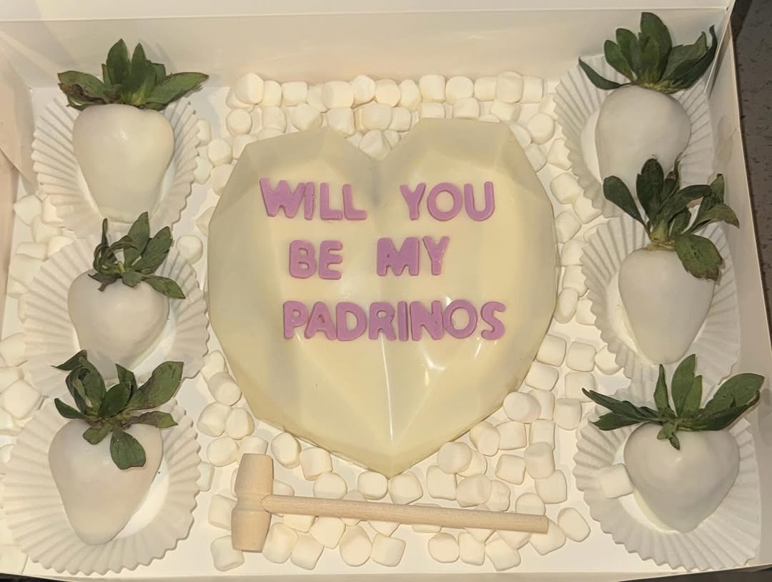 White chocolate breakable heart with WILL YOU BE MY PADRINOS in purple letters, six white chocolate strawberries, and a wooden mallet for a godparent proposal.