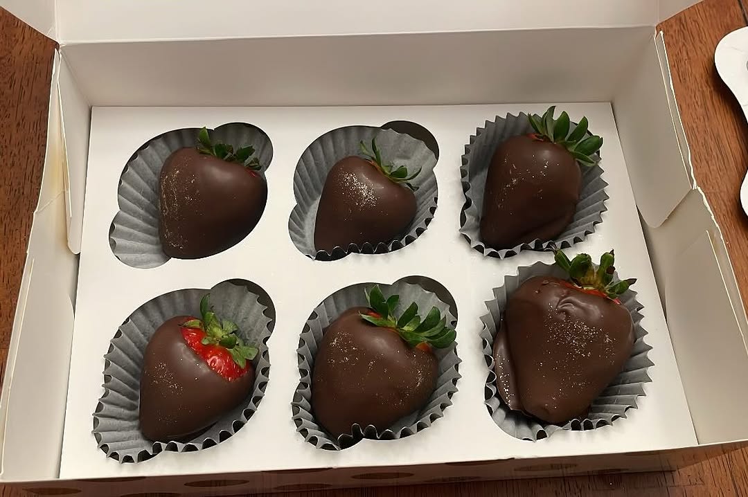 Half-dozen dark chocolate-covered strawberries with subtle gold shimmer dust, presented in a white bakery box with blue-gray paper liners.