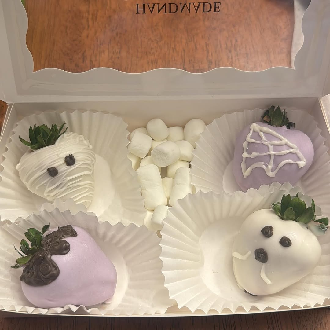 Four Halloween-themed strawberries in pastel lavender and white, featuring mummy, spiderweb, spider, and ghost designs with mini marshmallows.