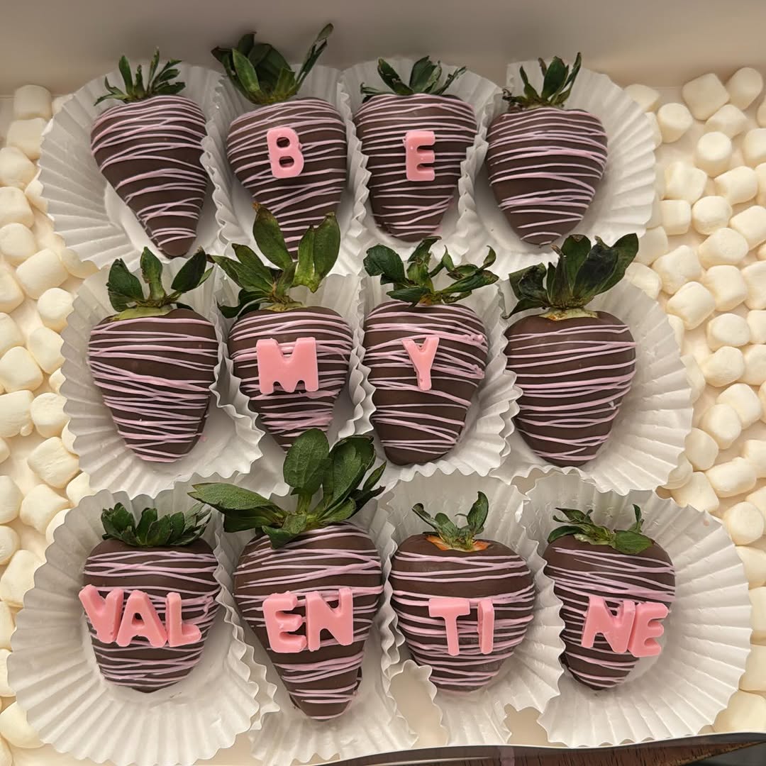 A dozen dark chocolate strawberries with pink drizzle and pink fondant letters spelling BE MY VALENTINE across three rows in ruffled cupcake liners.